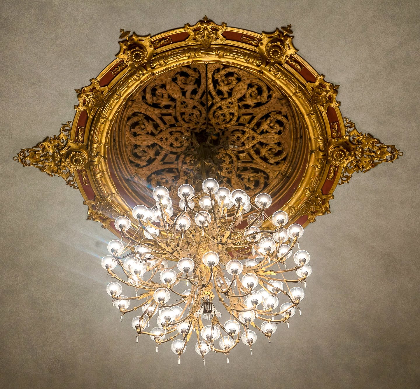 A large ornate chandelier with many bulbs hangs from a decorative golden ceiling medallion against a plain ceiling background.