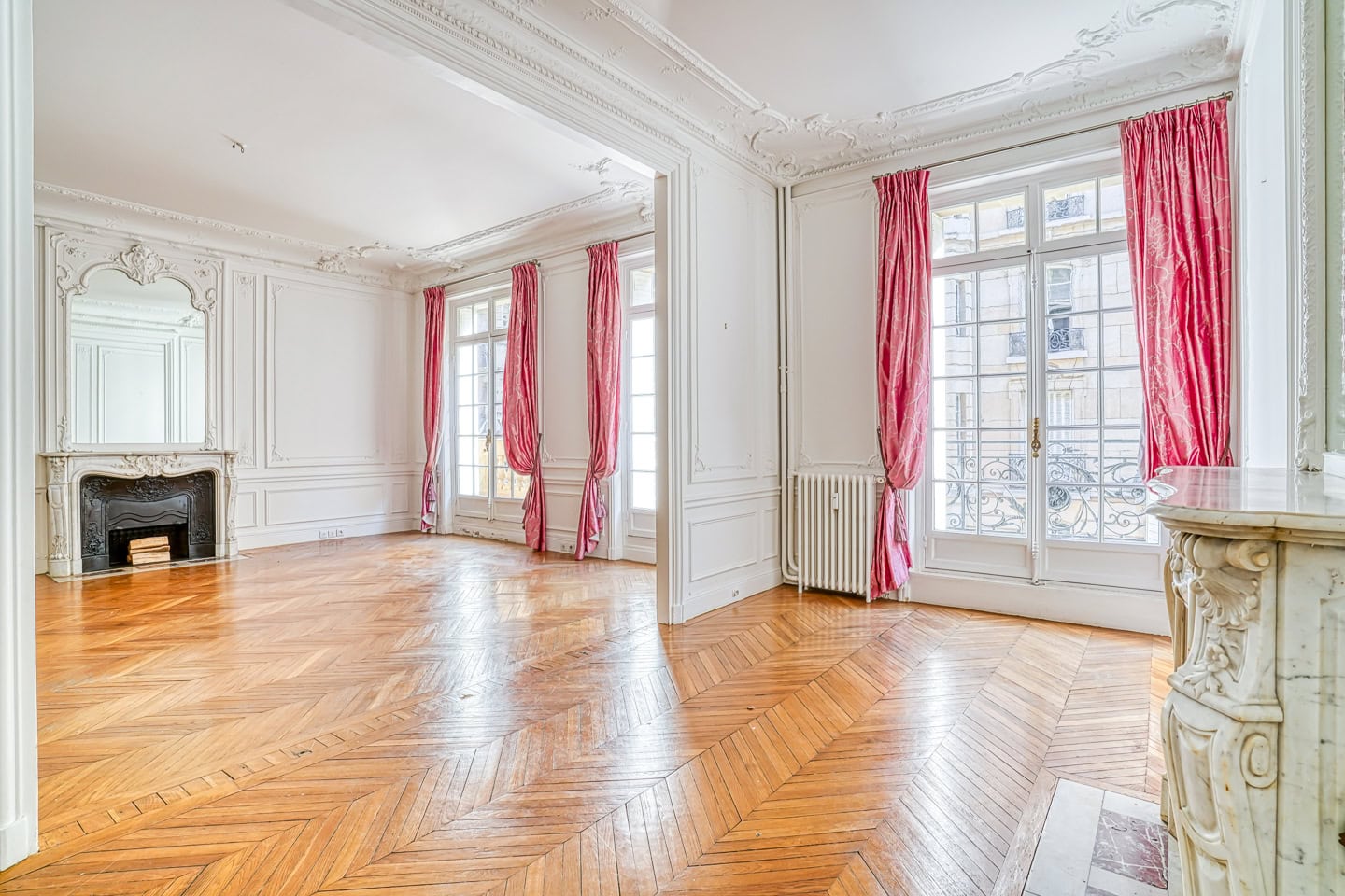 Spacious empty room with herringbone wood floors, ornate white walls, large windows with pink curtains, and a decorative fireplace.