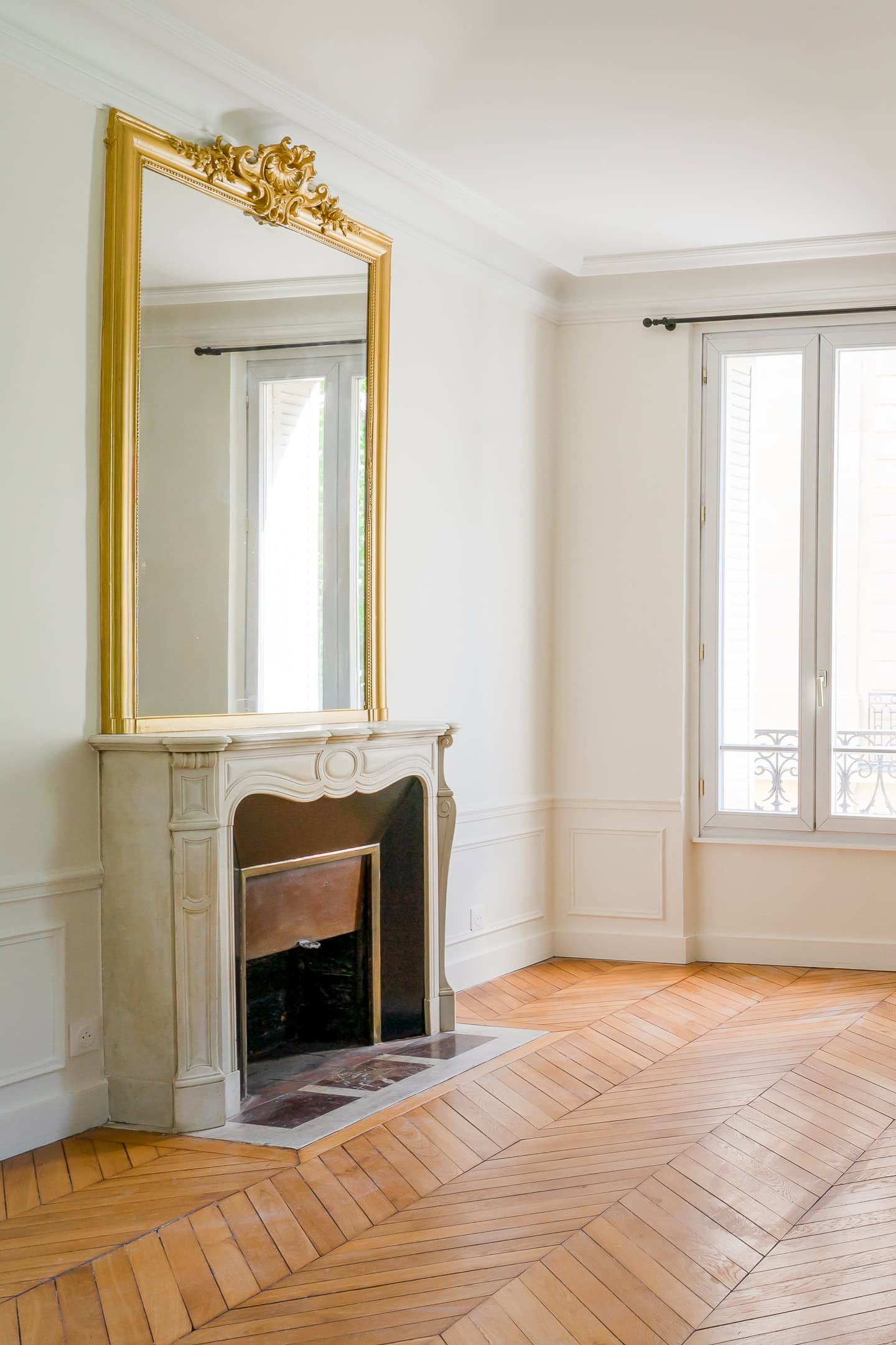 Bright room with a large window, decorative gold-framed mirror above a marble fireplace, and patterned wooden floor.