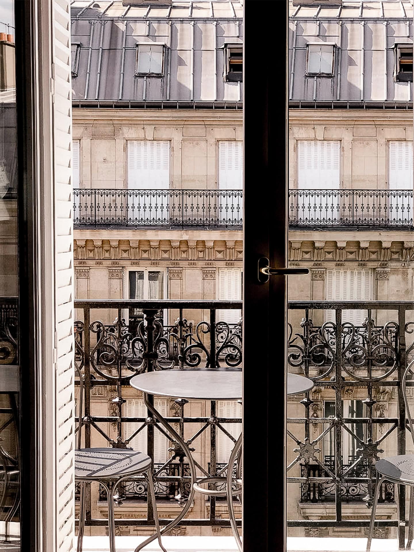 A small round table and two chairs on a balcony, seen through a glass door, with ornate railings and a building facade in the background.