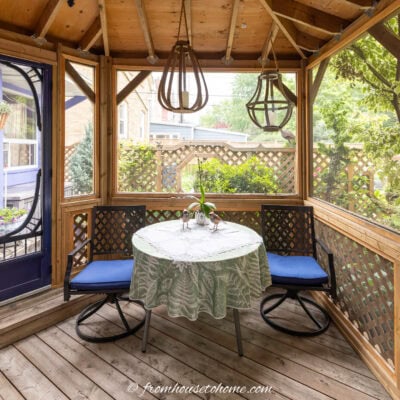 A small dining table with a green and white table cloth, two chairs and 2 hanging lanterns in an octagonal gazebo
