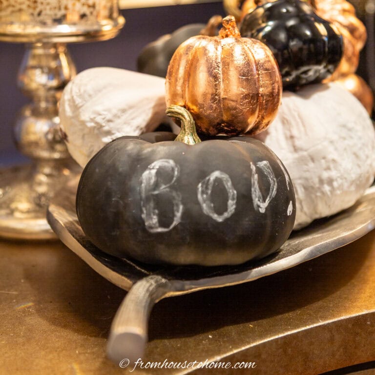 A black chalkboard pumpkin with the word "BOO" written on the front of it in a tray of black and white spray painted faux pumpkins with copper leaf pumpkins