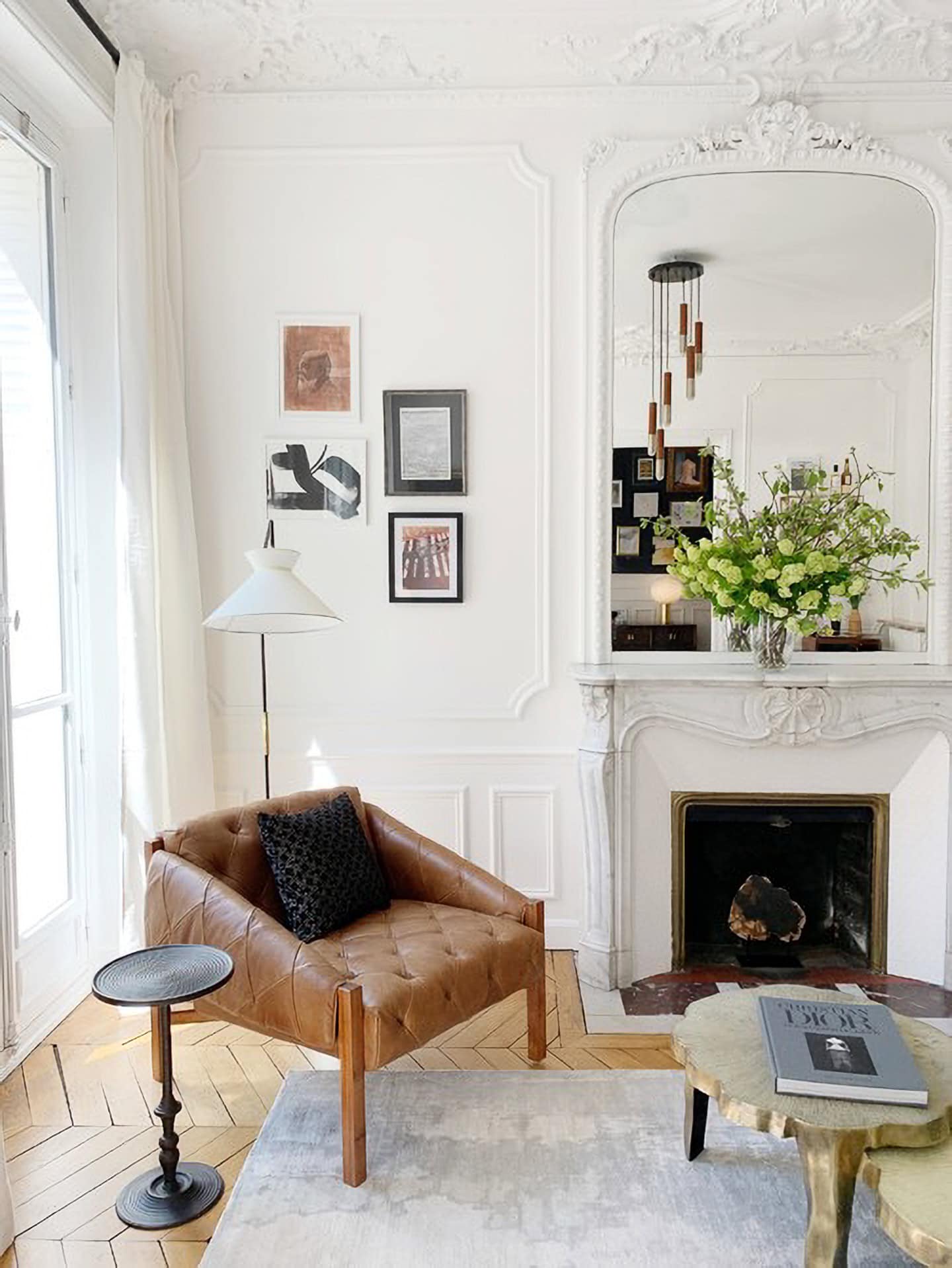 A stylish living room features a leather armchair, a marble fireplace with a mirror, a round table, and framed art on the white walls. A vase with flowers sits on the mantel.
