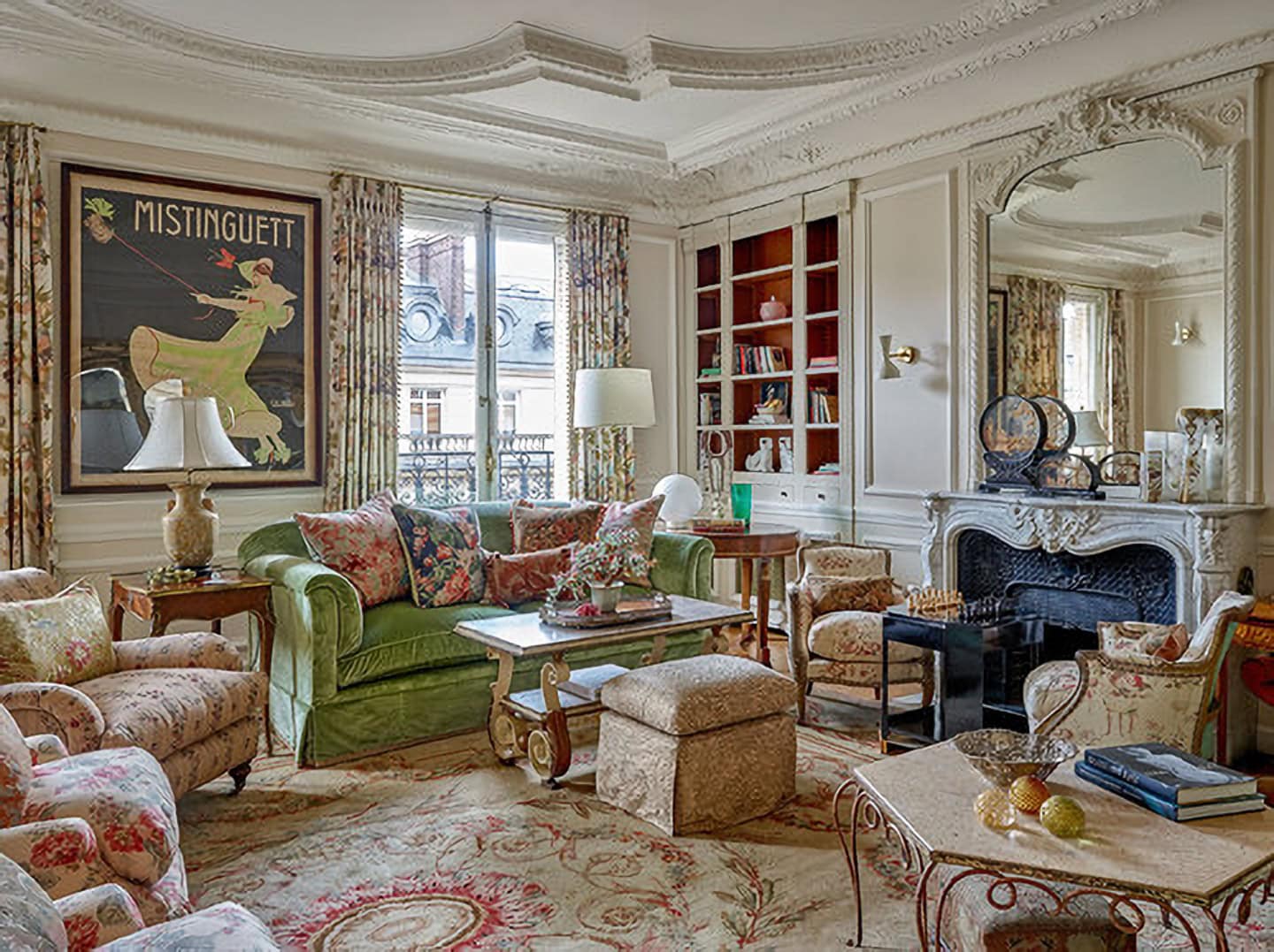 Elegant living room with floral furniture, ornate ceiling, large mirror above fireplace, bookshelves, a Mistinguett poster, and bright natural light from tall windows.