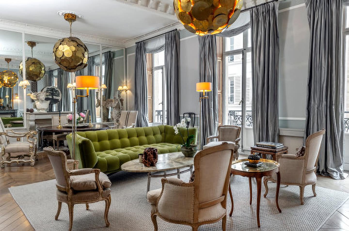 Elegant living room with a green velvet sofa, vintage armchairs, marble tables, tall windows with gray curtains, and gold pendant lights.