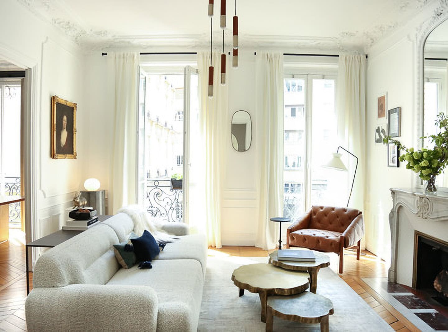 Bright living room with white walls and ornate molding, a modern white sofa, tufted leather chair, nested coffee tables, and a fireplace with a mirror and vase of flowers.