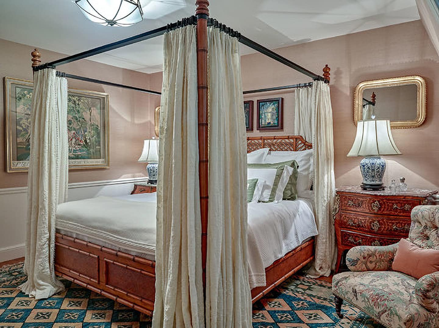 A four-poster bed with white curtains in a traditionally decorated bedroom featuring patterned carpet, lamps, artwork, and ornate furniture.