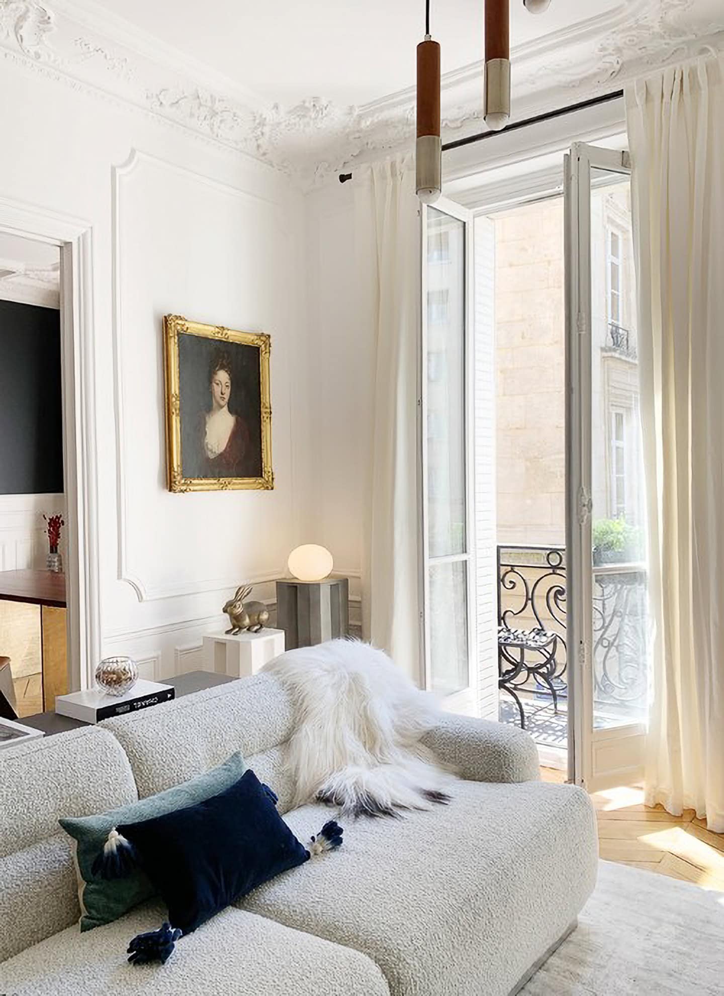 Bright living room with a white sofa, navy and white pillows, fur throw, framed portrait, modern lamp, and open doors leading to a balcony with ornate railing.