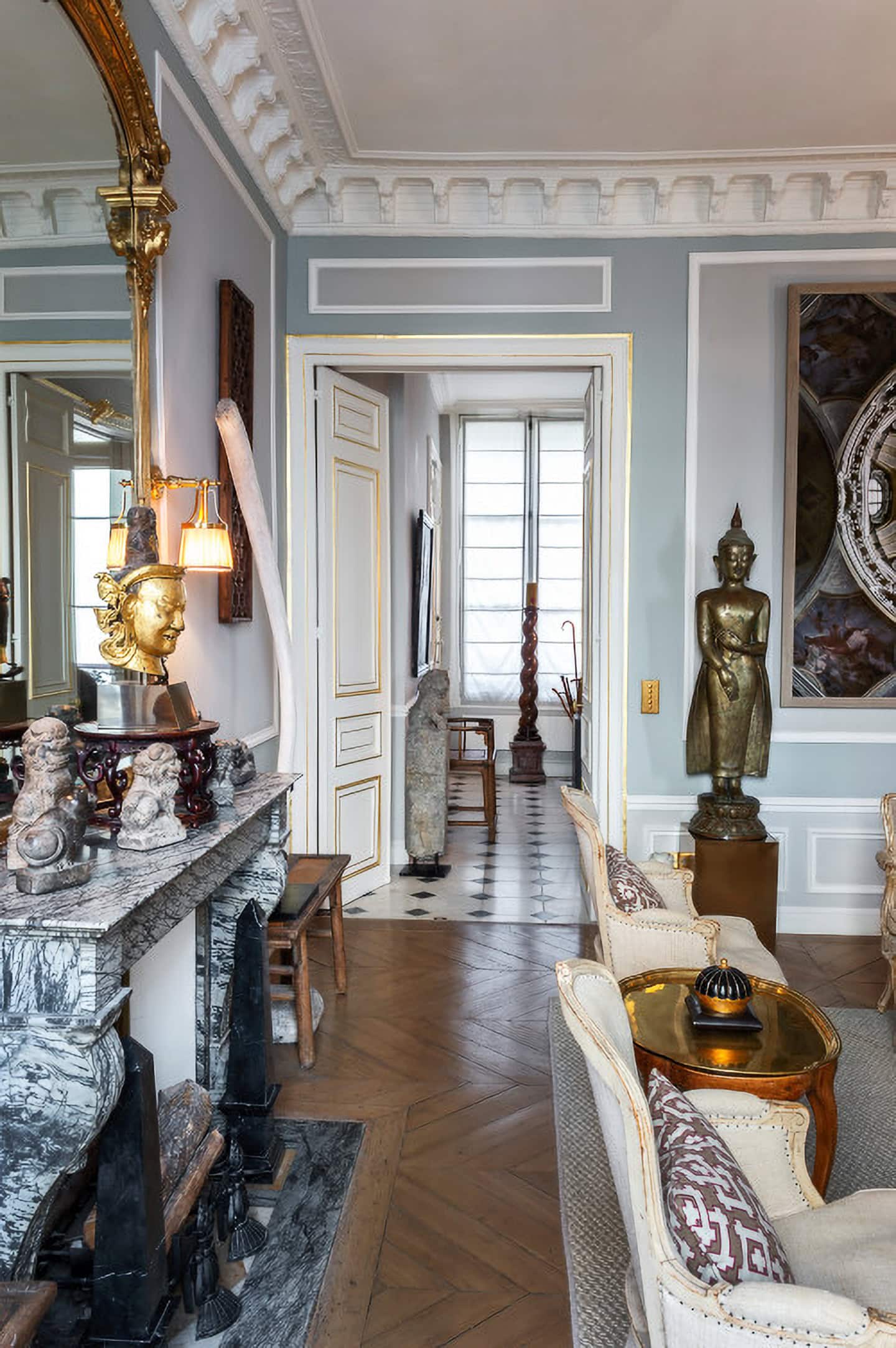 Eclectic living room with ornate fireplace, sculptures, gold-framed mirror, and classical decor elements; view into a hallway with additional statues and natural light from tall windows.