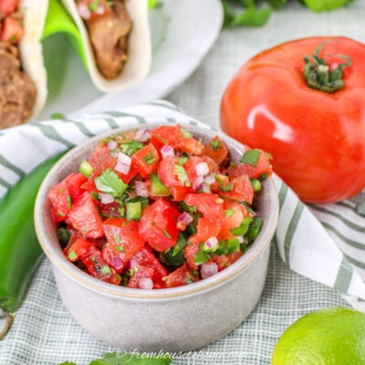 A bowl of fresh pico de gallo with diced tomatoes, onions, and jalapeños sits on a striped cloth next to a whole tomato, jalapeño, and lime. Tacos are partially visible in the background.