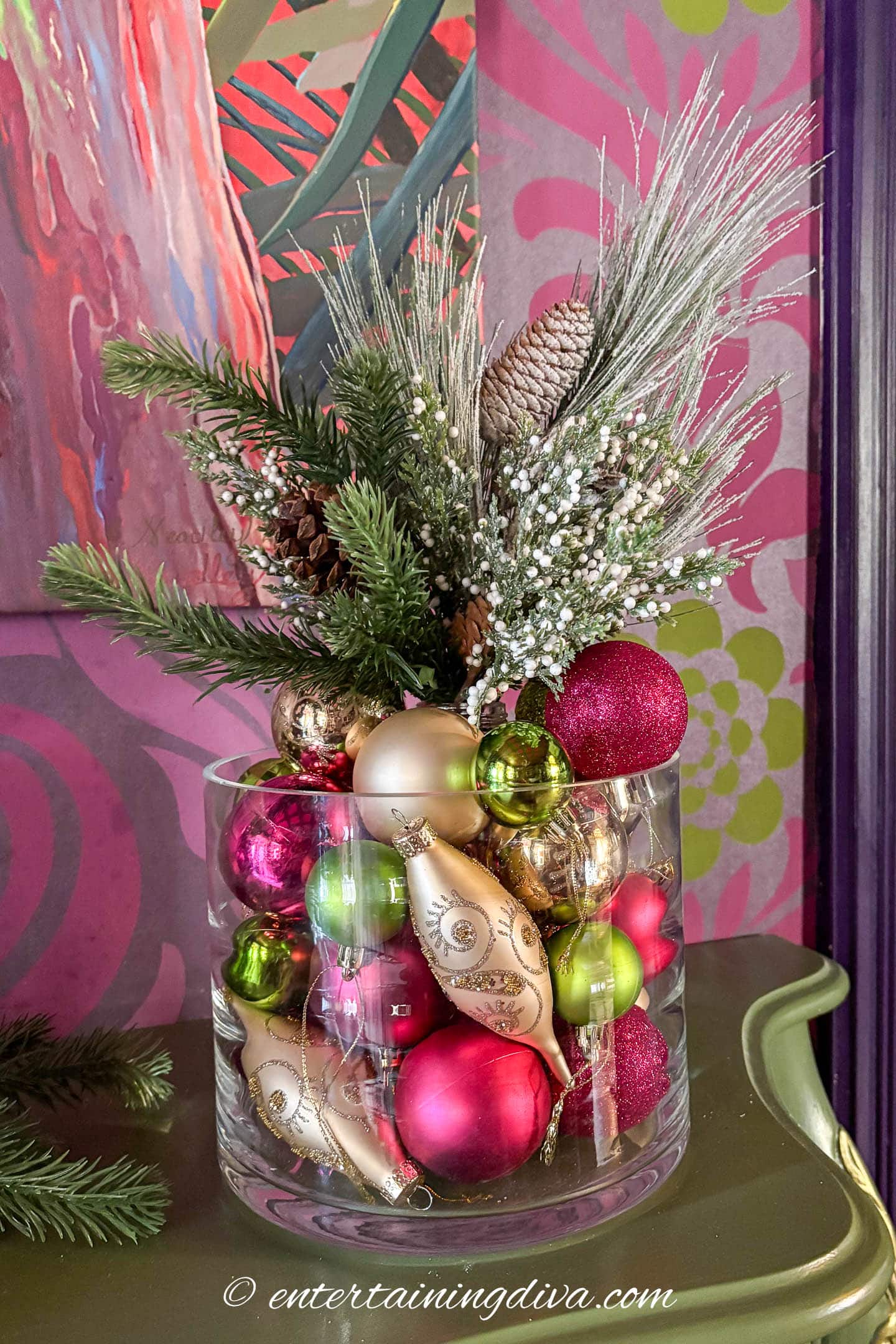 A clear glass vase filled with colorful Christmas ornaments, artificial pine branches, pinecones, and white berries, set on a green surface against a vibrant pink patterned background.