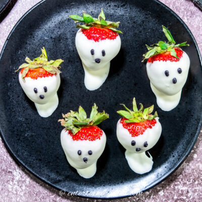 Five strawberries dipped in white chocolate to resemble ghosts, with chocolate dots for eyes and mouths, arranged on a black plate.